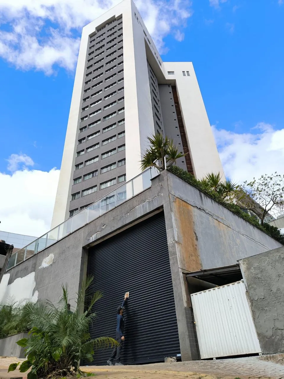 Edifício residencial alto com portão de enrolar na entrada da garagem e uma pessoa ao lado, mostrando a escala