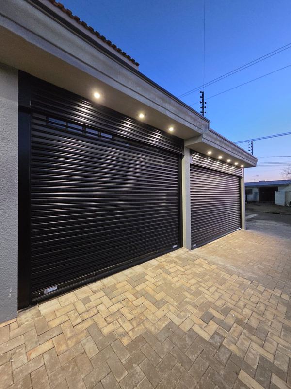 Portão de enrolar preto em duas garagens lado a lado, com iluminação embutida no beiral ao entardecer