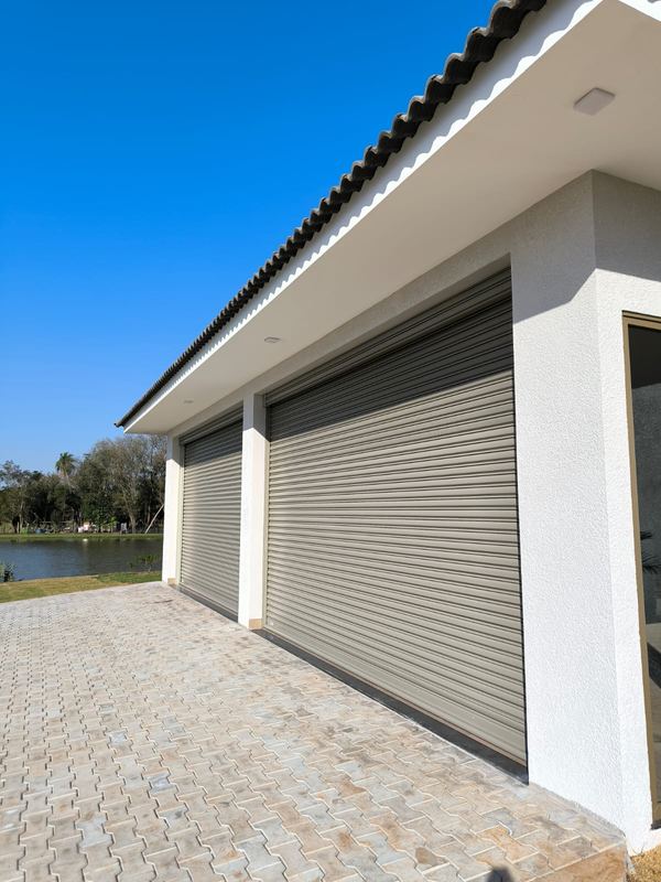 Portão de enrolar cinza em duas garagens de uma casa térrea, com um lago ao fundo